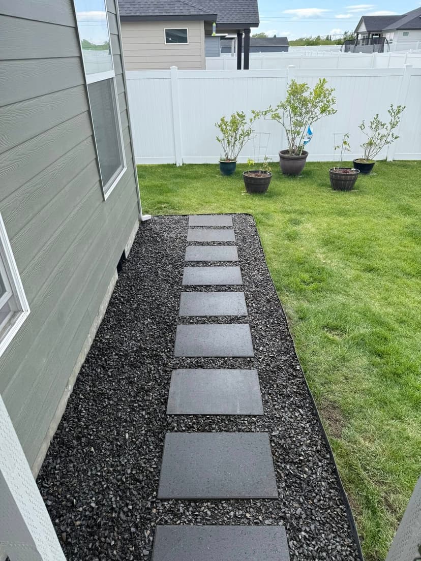 Stone pathway leading through gravel in a backyard with greenery and a white fence.